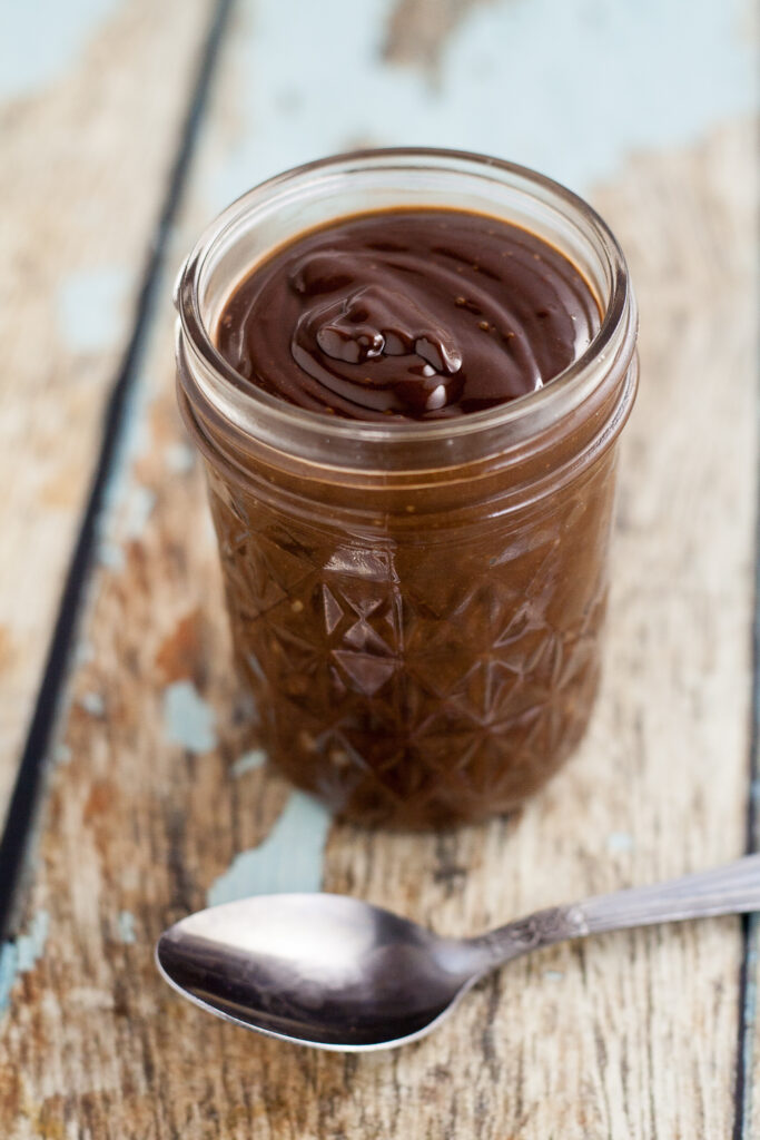 A quilted mason jar filled with chocolate ganache with a spoon in front on a rustic wood background.