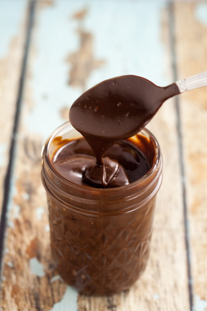 A spoon drizzling chocolate ganache into a quilted mason jar on a rustic wood background.