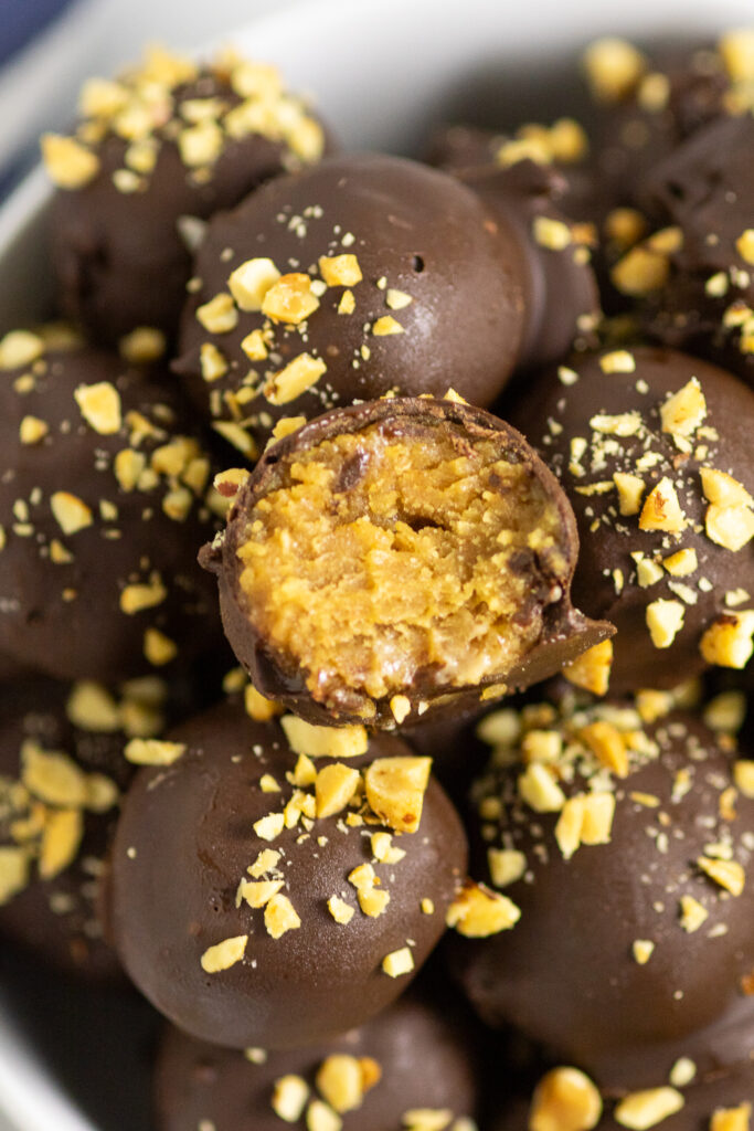 A pile of Nutter Butter Balls coated in chocolate and topped with chopped peanuts, and the top ball has a bite taken out showing the peanut butter ball center.