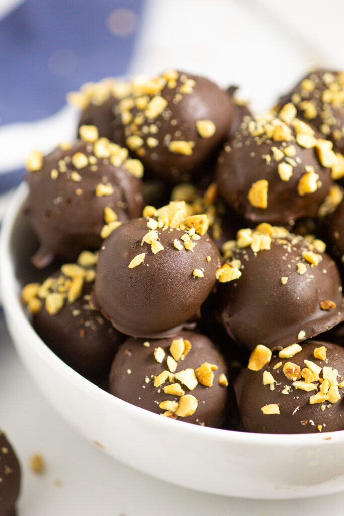 A stack of chocolate-dipped Nutter Butter Balls topped with chopped peanuts in a white bowl with a dark blue and white linen napkin behind on a white background.