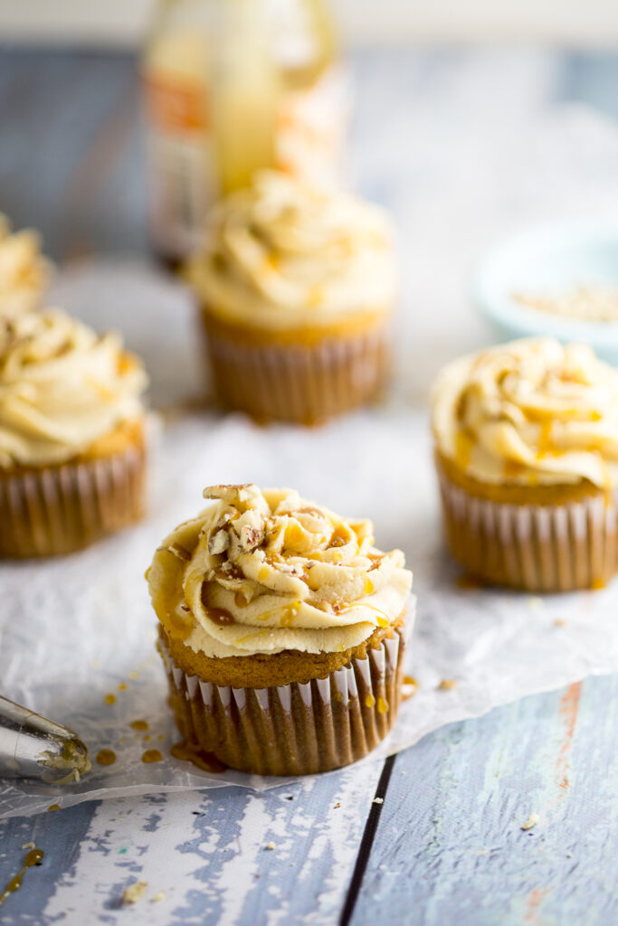 Sweet potato pecan cupcake on parchment paper and a rustic wood background topped with a caramel drizzle and chopped pecans with more cupcakes in the background.