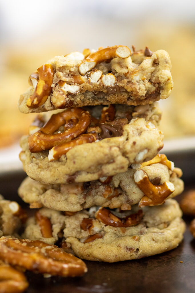 Stack of four toffee pretzel cookies surrounded by pretzel twists with the baking sheet full of cookies in the background. The top cookie has a bite taken out.