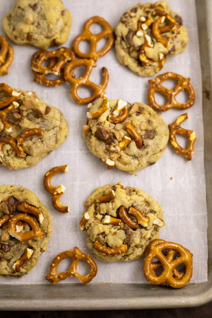 Overhead view of a baking sheet lined with parchment paper with Toffee pretzel cookies and pretzels twists scattered on it.