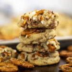 Stack of four toffee pretzel cookies surrounded by pretzel twists with the baking sheet full of cookies in the background. The top cookie has a bite taken out.