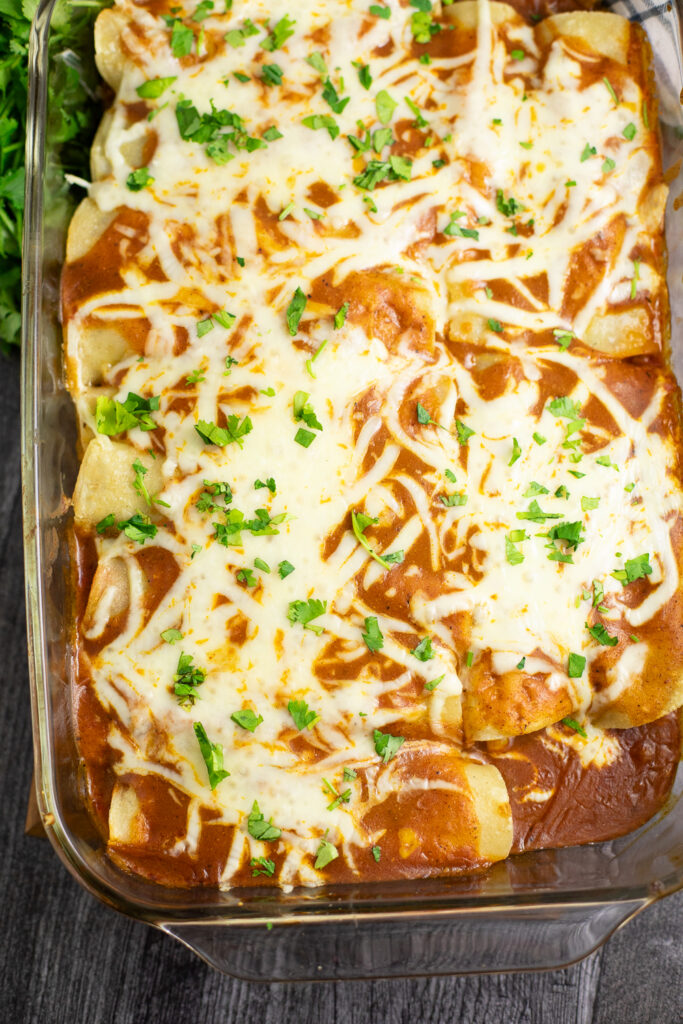 Overhead view of 3 bean enchiladas topped with red enchilada sauce, melted cheese, and fresh cilantro in a casserole dish on a rustic background.
