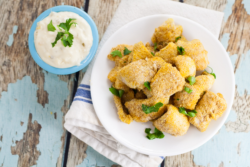 Cornmeal coated baked catfish nuggets on a white plate and tartar sauce in a small blue bowl on a rustic blue wood background.