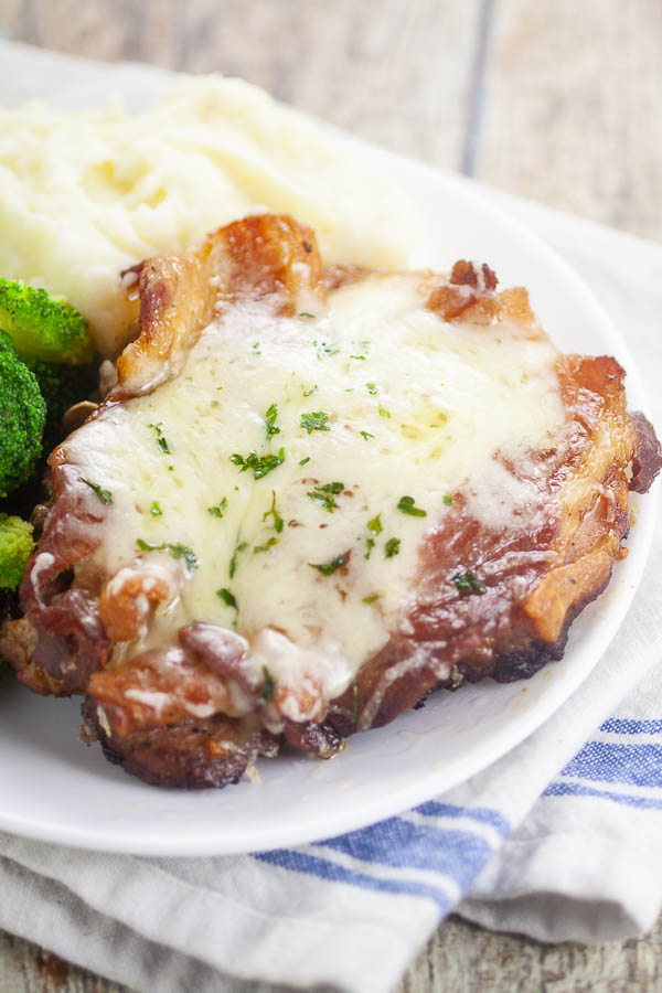 Thick cut pork chops with bacon and swiss cheese on white plate with veggies and potatoes.