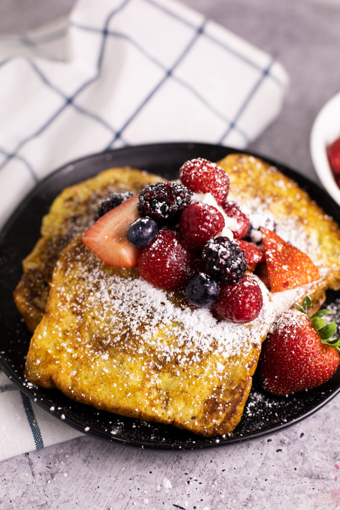 Three pieces of cinnamon French toast on a black plate topped with strawberries, blueberries, blackberries, raspberries, and powdered sugar.