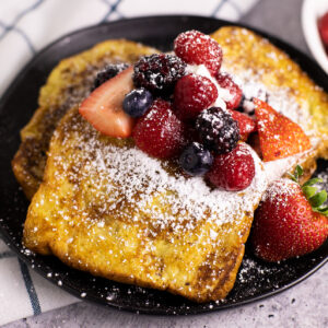 Three pieces of cinnamon French toast on a black plate topped with strawberries, blueberries, blackberries, raspberries, and powdered sugar.