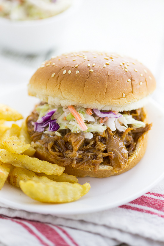 Easy Slow Cooker BBQ Pulled Pork topped with homemade coleslaw on a sesame seed bun next to crinkle cut fries on a small plate