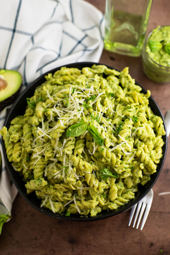 Overhead view of a bowl of avocado pesto pasta topped with fresh basil and grated parmesan and surrounded by a halved avocado, fork, linen napkin, small bowl of avocado pesto, and an oil bottle.
