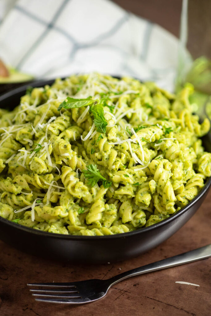 Side view of a bowl of avocado pesto pasta topped with grated parmesan and fresh basil with a fork in front and an oil bottle and linen napkin behind.