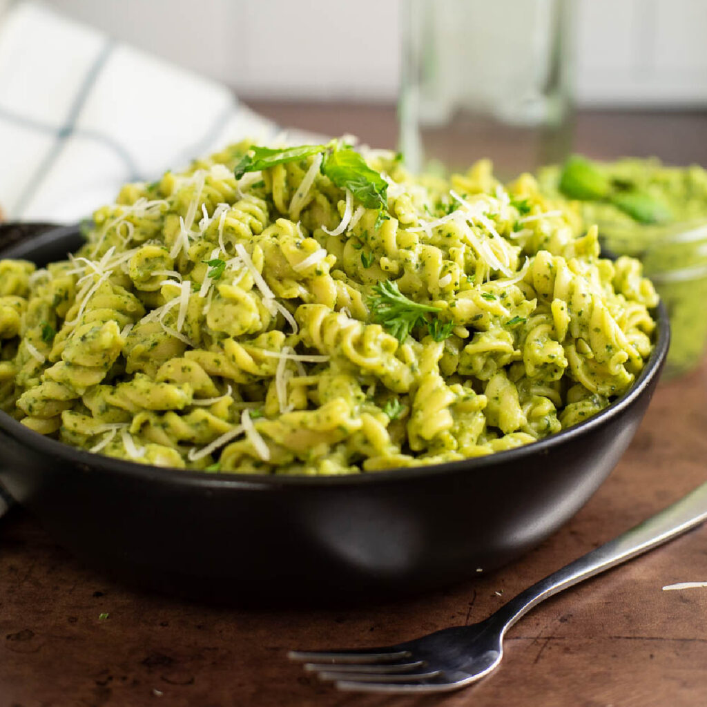 Side view of a bowl of avocado pesto pasta topped with grated parmesan and fresh basil with a fork in front and an oil bottle and linen napkin behind.
