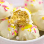 A lemon Oreo truffle with a bite taken out sitting on top of a bowl of other truffles, all coated in white chocolate and topped with pink and yellow sprinkles.