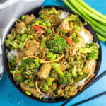 Overhead view of easy chicken stir fry on white rice in a black matte bowl next to black chop sticks, green onions, and a linen napkin on a bright blue background.