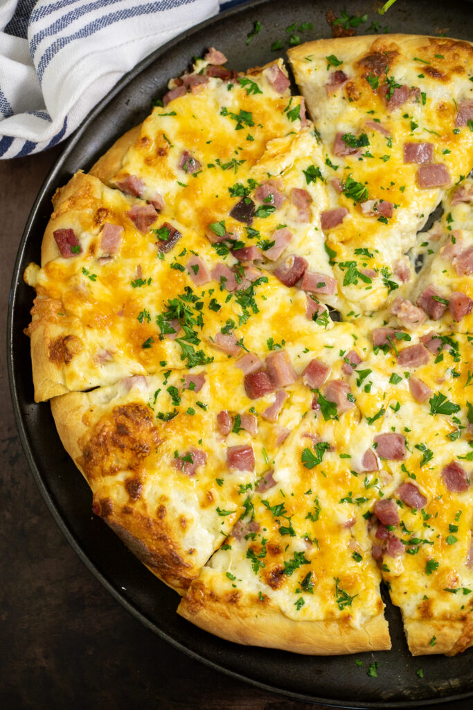 Overhead view of half of a homemade ham and cheese pizza on a pizza sheet topped with gooey cheese sauce, melted cheese, cubed ham, and parsley. The pizza is cut into triangle slices and is next to a striped linen.