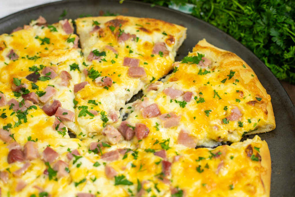 Close up of a slice of ham and cheese pizza topped with gooey cheese, diced ham, and parsley.