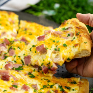 A picture of a piece of ham and cheese pizza being lifted by a hand from the whole pizza with melty cheesy pulling from it and fresh parsley behind.