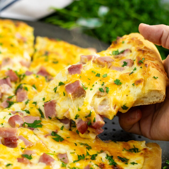 A picture of a piece of ham and cheese pizza being lifted by a hand from the whole pizza with melty cheesy pulling from it and fresh parsley behind.