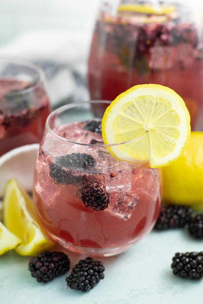 Blackberry Lemon Spritzer in a stemless wine glass with ice and blackberries and a lemon slice on the rim. There's lemon wedges and blackberries around the glass and a pitcher behind.