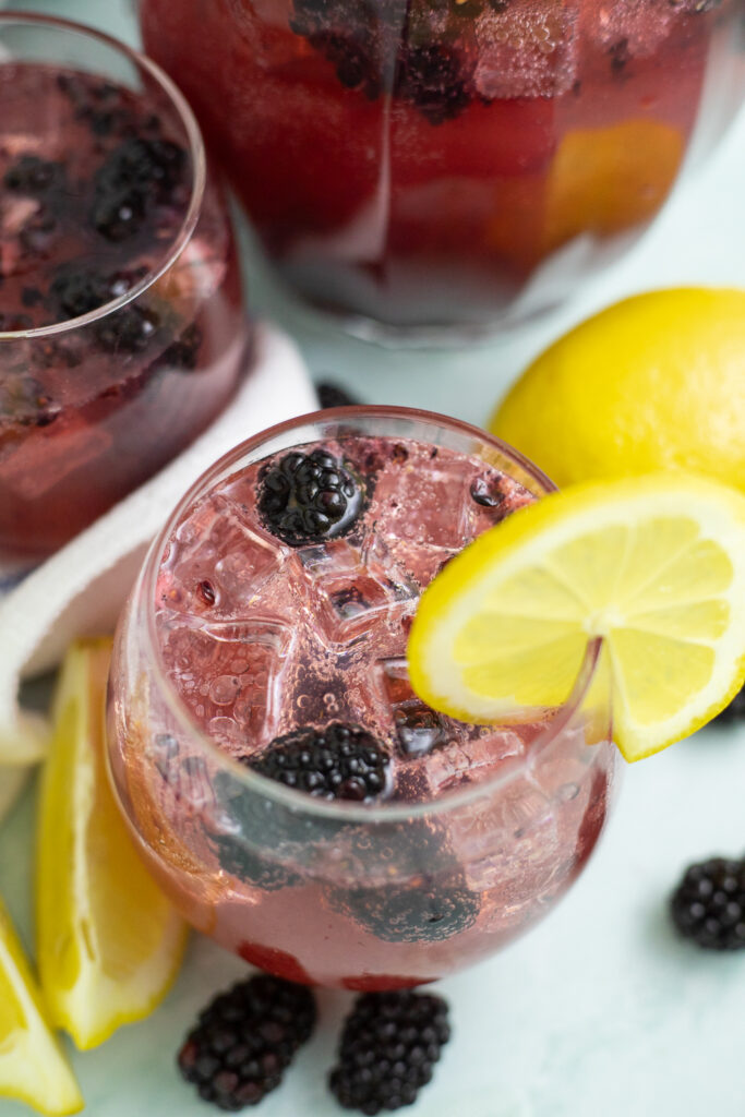 Overhead view of a Blackberry Lemon Spritzer in a stemless wine glass with ice and blackberries and a lemon slice on the rim. There's lemon wedges and blackberries around the glass and a pitcher behind.