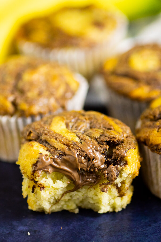 Nutella banana muffins with a bite taken out with more muffins behind on a dark navy background.