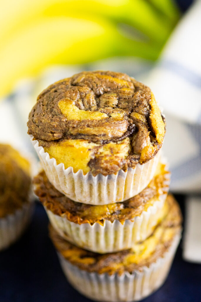 Three Nutella banana muffins in cupcake wrappers stacked on top of each other with a linen napkin and fresh bananas behind.