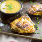 Grilled chicken thigh on a baking sheet with more chicken behind it, a small bowl of maple dijon sauce, and fresh chives scattered around.