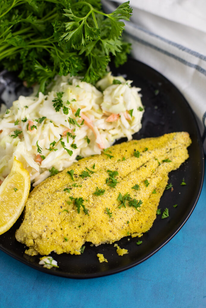 Lemon Pepper Baked Catfish on a black plate with a lemon wedge and creamy coleslaw.