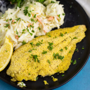 Lemon Pepper Baked Catfish on a black plate with a lemon wedge and creamy coleslaw.