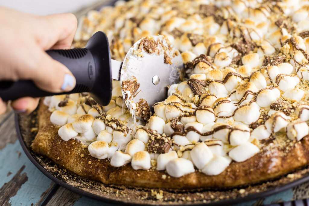 A slice of s'mores pizza being cut with a pizza cutter out of the full pizza