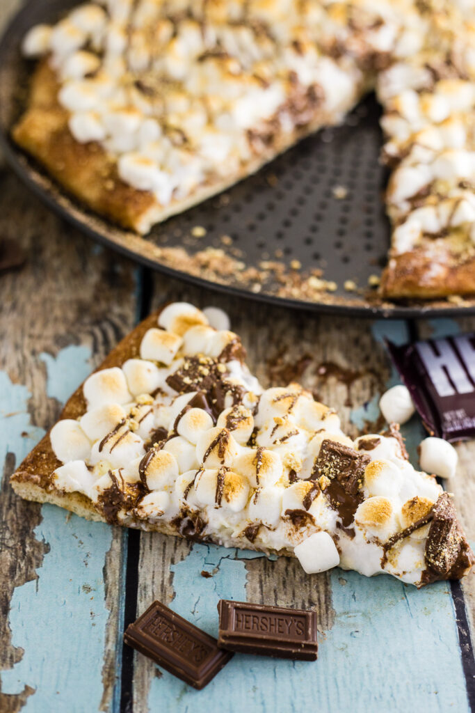 A slice of s'mores pizza on a rustic wood background sitting in front of the full pizza 