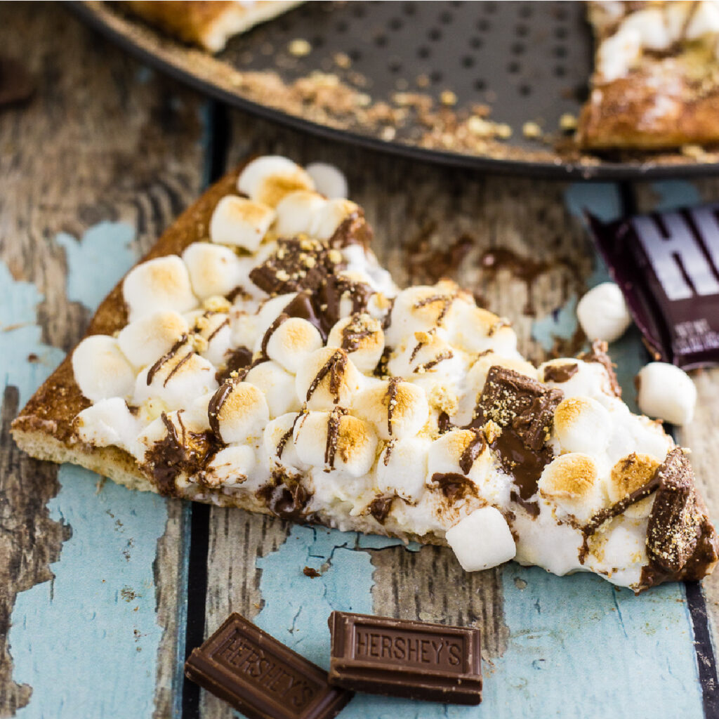A slice of s'mores pizza on a rustic wood background sitting in front of the full pizza