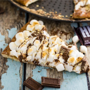 A slice of s'mores pizza on a rustic wood background sitting in front of the full pizza