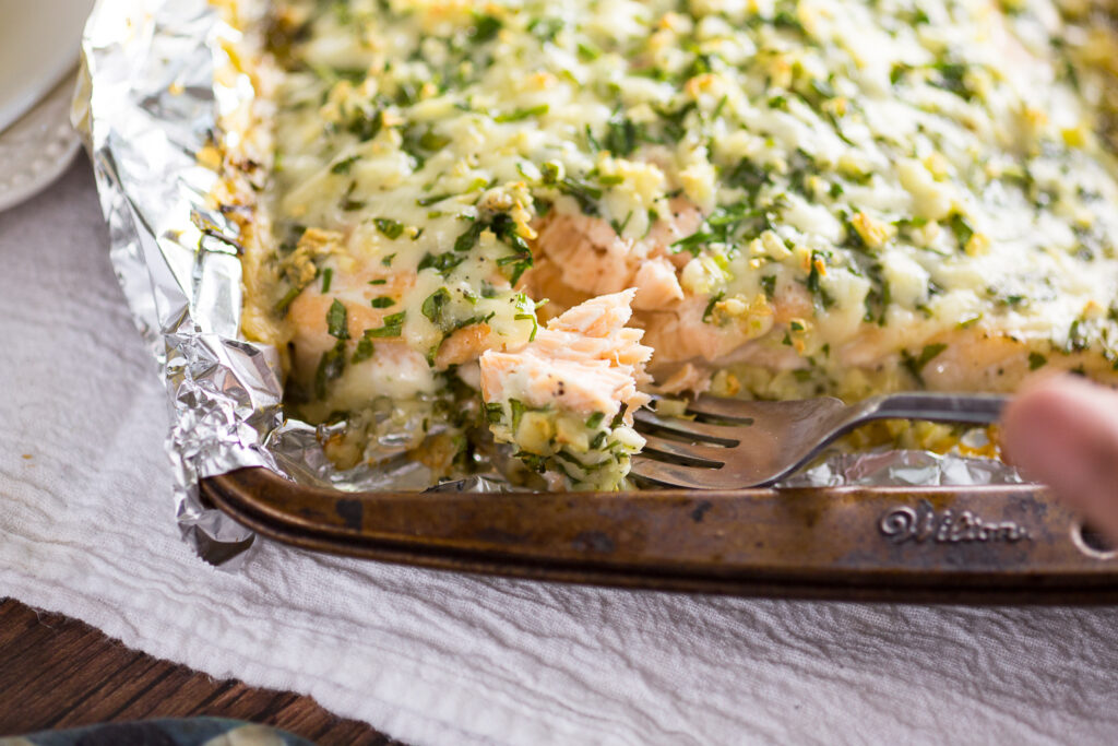 A fork taking a bite out of parmesan garlic herb salmon on a baking sheet.
