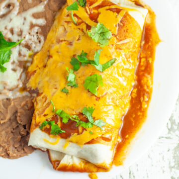 Overhead view of a rice and black bean burrito topped with red enchilada sauce, melted cheese, and fresh cilantro, next to refried beans on a white plate.