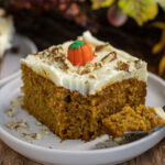 Pumpkin sheet cake topped with chopped pecans and a creme pumpkin. The cake has a bite taken out sitting on a fork on a small plate.