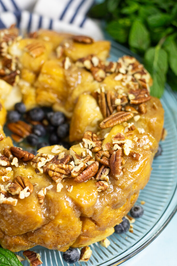 Close up view of half of a bundt-shaped caramel pull apart cake topped with chopped pecans on a glass plate with some fresh blueberries. The plate is on a bright blue background next to some fresh mint.