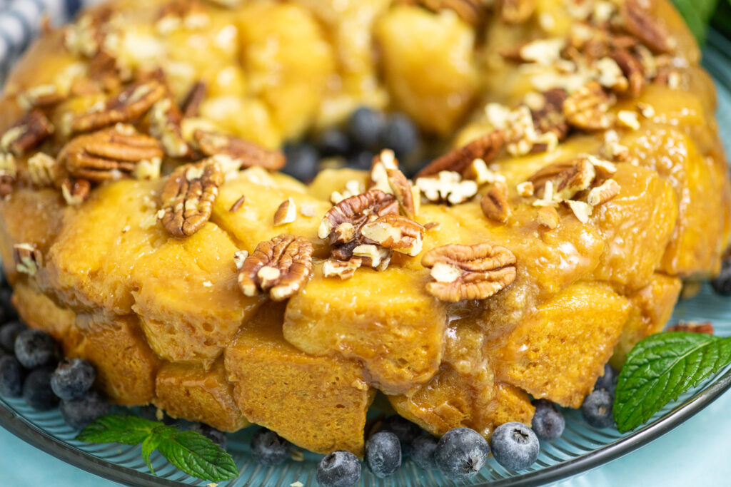 Side view of a caramel pull apart cake topped with chopped pecans and next to fresh blueberries and mint leaves on a glass plate on a bright blue background.