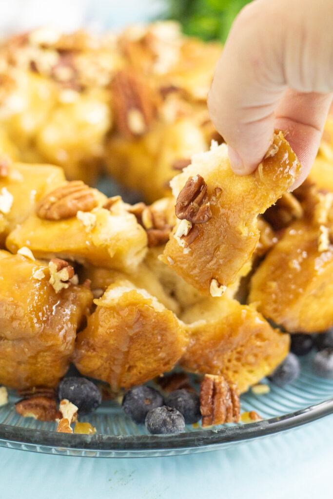 A hand pulling a piece of caramel pull apart cake from the full cake on a glass plate with a bright blue background.