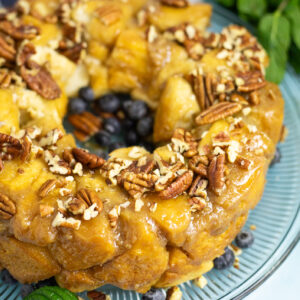 Close up view of three-quarters of a bundt-shaped caramel pull apart cake topped with chopped pecans on a glass plate with some fresh blueberries. The plate is on a bright blue background next to some fresh mint.