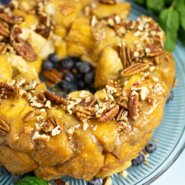 Close up view of three-quarters of a bundt-shaped caramel pull apart cake topped with chopped pecans on a glass plate with some fresh blueberries. The plate is on a bright blue background next to some fresh mint.