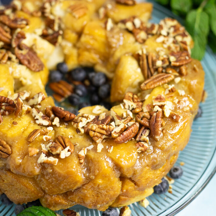 Close up view of three-quarters of a bundt-shaped caramel pull apart cake topped with chopped pecans on a glass plate with some fresh blueberries. The plate is on a bright blue background next to some fresh mint.
