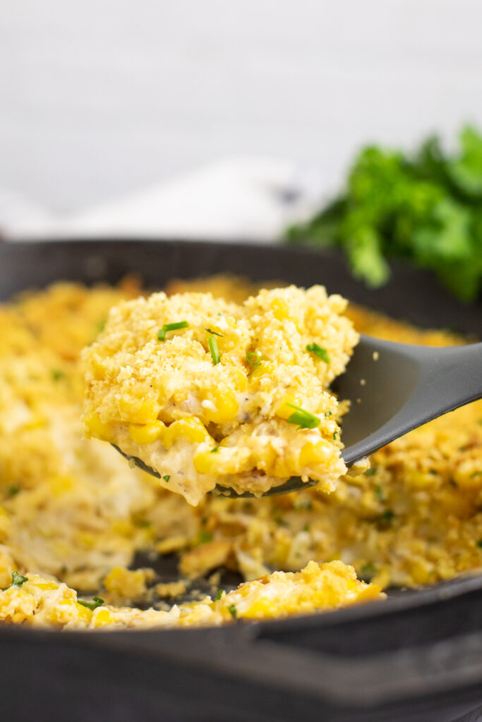 Silicone spoon scooping a large portion of cheesy corn bake topped with buttery cracker topping out of a cast iron skillet.