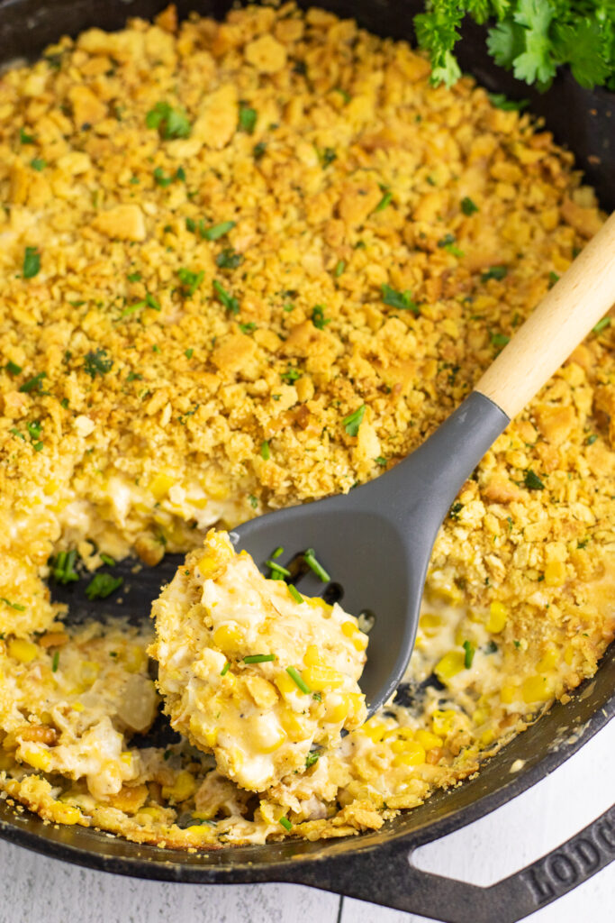 Overhead view of cheesy corn bake in a cast iron skillet topped with buttery cracker topping and fresh chives and a spoon in the middle.