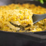Silicone spoon scooping a large portion of cheesy corn bake topped with buttery cracker topping out of a cast iron skillet.