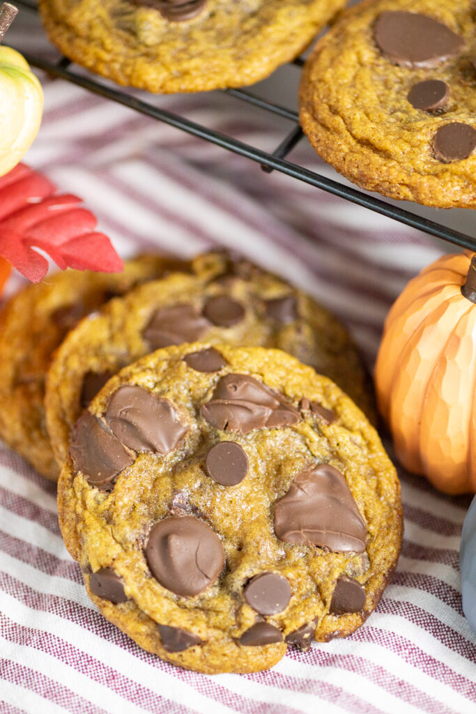Two pumpkin chocolate chip cookies sitting on a red and white striped linen napkin next to mini pumpkins with a cooling rack full of cookies behind.