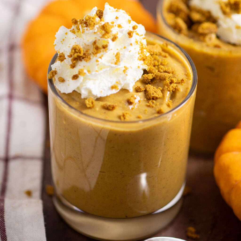 Pumpkin pudding in a small glass topped with whipped cream and gingersnap crumbs with another glass of pudding behind. They're surrounded by mini pumpkins and a linen napkins.