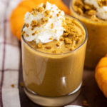 Pumpkin pudding in a small glass topped with whipped cream and gingersnap crumbs with another glass of pudding behind. They're surrounded by mini pumpkins and a linen napkins.