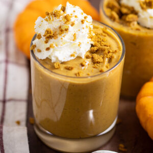 Pumpkin pudding in a small glass topped with whipped cream and gingersnap crumbs with another glass of pudding behind. They're surrounded by mini pumpkins and a linen napkins.